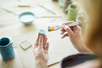 Woman with paintbrush painting handmade clay dice in blue color over workplace