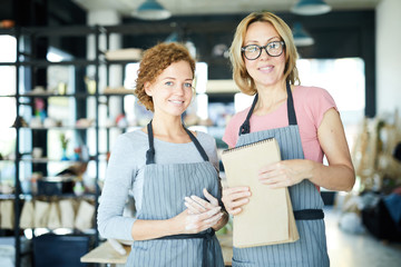 Two happy young creative females in aprons looking at you in workshop