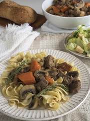 Plate of beef burgundy served over egg noodles, with a side of tossed green salad and baked bread loaf. A serving bowl of beef burgundy is in the background