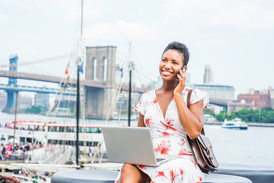 African American Woman With Short Afro Hair Traveling In New York, Sitting By East River, Talking On Phone, Working On Laptop Computer In Same Time. Manhattan, Brooklyn Bridges, Boat On Background..