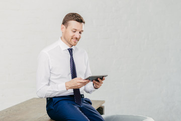Positive male financier wears white shirt, tie, sits on table, installs new application on touch pad, connected to wireless internet. Successful businessman reads notification on modern device