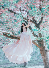 Obraz na płótnie Canvas Incredible, gentle elf in a luxurious, gently pink dress that waving in the wind. The princess with long curly hair sits on a branch of a blooming tree, like a bird. Vanilla color art photo