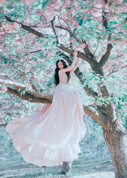 Incredible, gentle elf in a luxurious, gently pink dress that waving in the wind. The princess with long curly hair sits on a branch of a blooming tree, like a bird. Vanilla color art photo