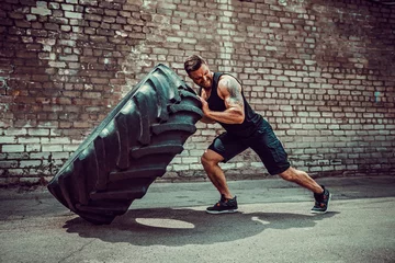 Gordijnen Muscular bearded tattooed fitness man moving large tire in street gym. Concept lifting, workout training. © zamuruev