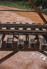 Train tracks on a wooden and steel brown bridge over an orange muddy river in Peru
