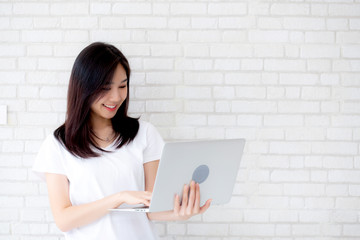 Beautiful of portrait young asian woman smiling and standing holding laptop on brick cement wall background, girl working computer, business and lifestyle concept.