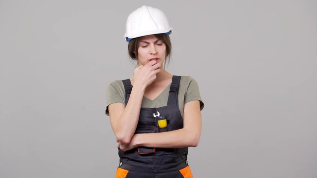 Portrait of brooding woman builder in helmet and jumpsuit touching chin and thinking while working on factory or manufacture, isolated over gray background