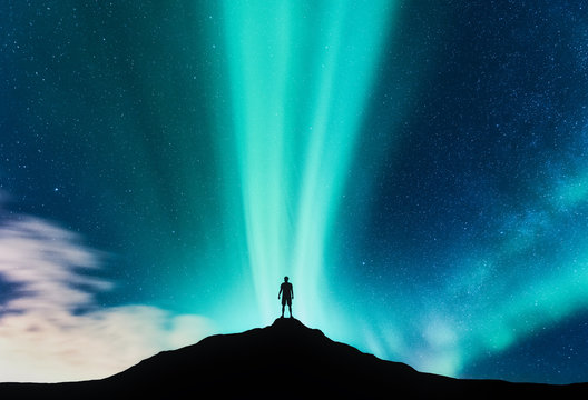 Aurora borealis and silhouette of standing man on the mountain. Aurora and traveller. Starry sky and green polar lights. Night landscape with northern lights and people. Concept. Nature background
