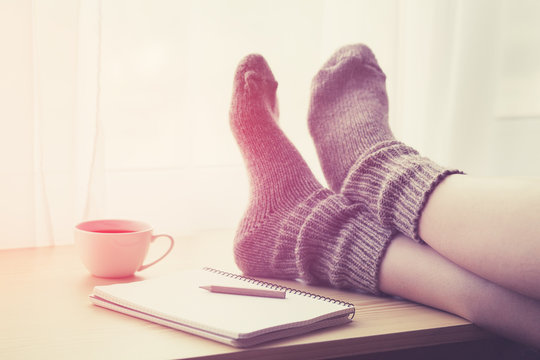 Woman resting keeping legs in warm socks on table with morning coffee and notebook