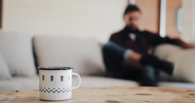 Attractive bearded man with smartphone sitting on sofa at modern loft home in the living room and chatting with friends. In the foreground there is a cup with a hot drink.