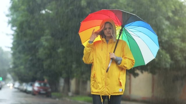 outside portrait of beautiful thoughtful woman in autumn yellow jaket holding colorful umbrella and walking on the street waiting for date rainy weather falling raindrops urban lifestyle fashion