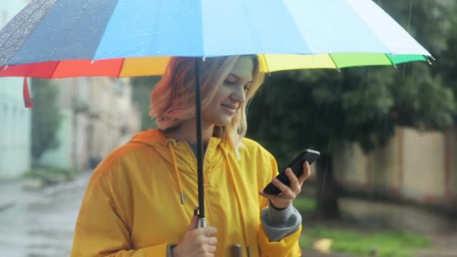 outdoors portrait of young blond woman with colorful umbrella using modern smartphone mobile while standing under the rain on the road waiting for taxi car with blur background busy city lifestyle