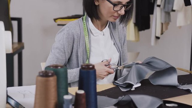 Close-up of a dressmaker that sits at the table and cuts gray material with scissors for sewing clothes in a small stylish atelier. Young woman tailor working with scissors at the studio with