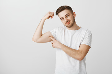 Wanna touch my strong biceps. Cute and flirty handsome male with beard in white casual t-shirt tilting head and smiling with delighted and pleased look poking himself in mascule on right hand