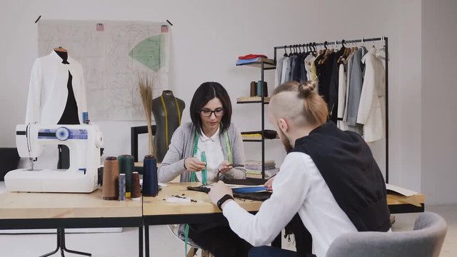 Two people fashion designers choosing fabric at the office with different tailoring tools and clothes. Young woman and man fashion designers or tailors working with fabrics sitting at the beautiful