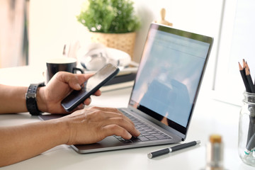 Cropped shot of businessman holding mobile phone while working with laptop computer at desk.