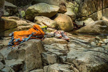 an orange backpack and two pair of shoes on the rock on the side of the creek.