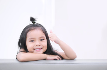 Asian children or kid girl smile and student thinking with show thumb finger for good or like with new idea and happy fun on sofa at kindergarten nursery and child hospital or school with brown space