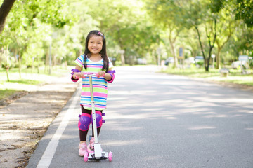 asian children or kid girl smile happy with fun and play or ride scooter for exercise with healthy and balance learn on holiday at public park or green garden and wear knee elbow protection for safety