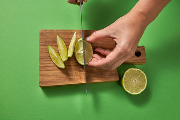 A girl's hands cut a green natural lime to slices with sharp knife on a wooden board on a green....