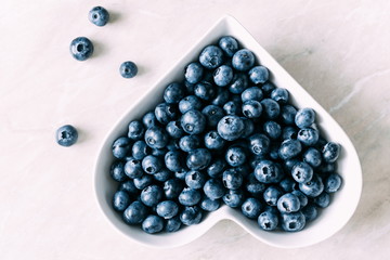 Blueberries berry in a plate in the shape of a heart on a light table. Flat lay, top view, copy space 