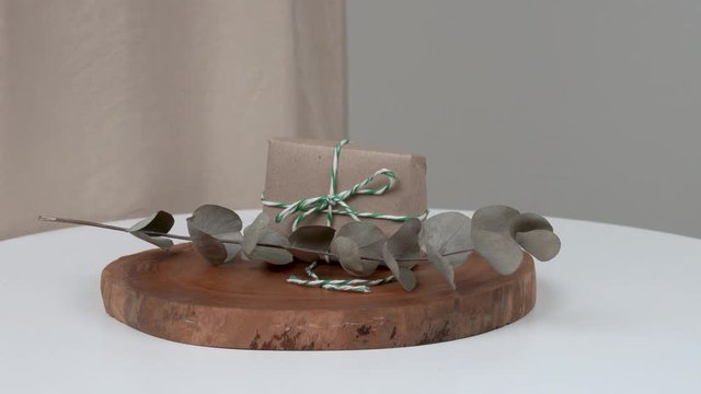 A close-up of a woman's hand holding a wrapped and tied bar of natural soap vertically. The bar is lying on a wooden board. She is putting an eucalyptus twig in front of the bar.