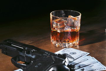 Glass of whiskey or cognac, gun, playing cards with money on the black mirror table. The concept of criminal.