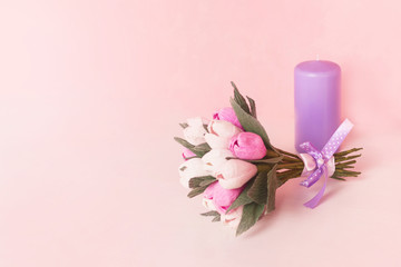 Bow of purple ribbon on a bouquet of paper flowers. On a background a candle. Flowers are made of paper.
