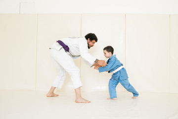 Father and little kid son are engaged in wrestling jiu-jitsu in the gym in a kimono. Trainer...