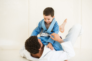 Father and little kid son are engaged in wrestling jiu-jitsu in the gym in a kimono. Trainer...