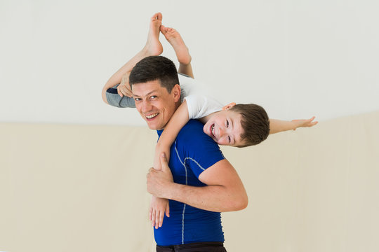 Dad And Son Training In Gym Together. Portrait Of Young Father With Little Kid Boy At Sport Training. Child Sits On Father Shoulders And Hugs His Neck, They Smiling And Laughing