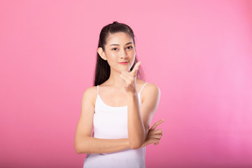 Portrait of a smiling attractive woman in white tanktop outfit with finger pointing pose while standing and smiling at camera isolated over pink background.
