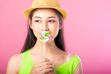 Portrait of a smiling attractive woman in summer outfit and hat posing with lollipop over her mouth and looking at camera isolated over pink background.