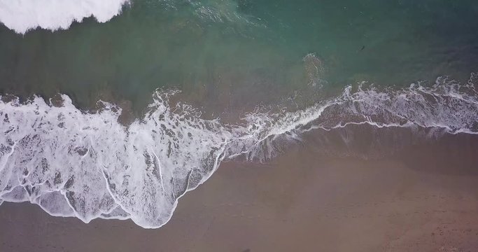 Aerial top view waves break on white sand beach at sunset. Sea waves on the beautiful beach aerial view drone 4k shot. Bird's eye view of ocean waves crashing against an empty beach from above.