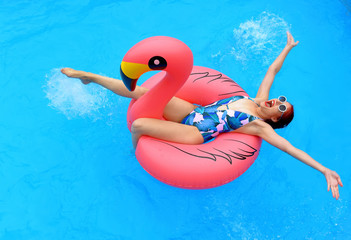 A young girl wearing a bathing suit and a white summer hat sits relaxed in an inflatable swim ring. She poses in  a pool smiling at the camera.