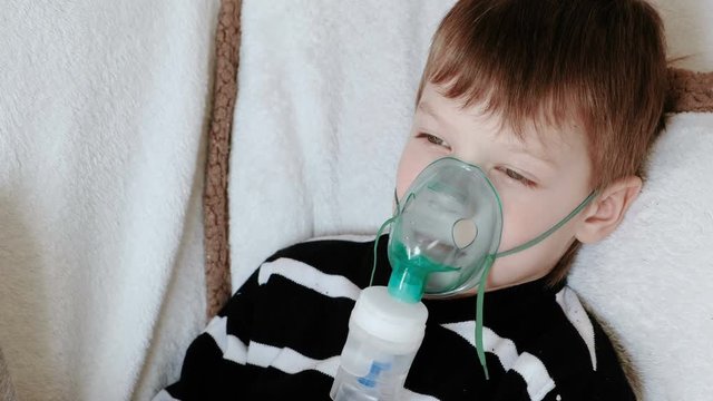 Using nebulizer and inhaler for the treatment. Boy inhaling through inhaler mask lying on the couch.