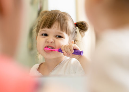 Cute little child girl brushing teeth and looking in mirror in bathroom