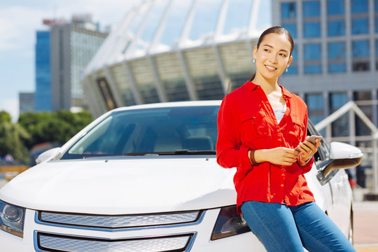 Digital age. Cheerful happy woman holding her smartphone while standing near the car