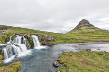 Landscape view of famous Kirkjufell Mountain and Waterfall landmark with long exposure smooth water wide angle with nobody, Grundarfjordur, green overcast cloudy day