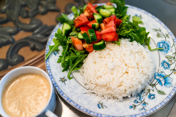 Closeup of plain white jasmine long grain cooked rice with arugula tomato salad on plate, cup with sauce or coffee drink, table healthy vegan diet food