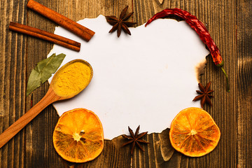 Piece of paper on wooden background. Spices, kitchen herbs lay around white paper. Cinnamon sticks, dried orange and star anise lay around blank paper for recipe, copy space. Culinary recipe concept