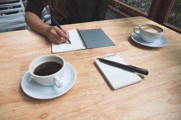 Closeup image of a woman's hand writing down on a white blank notebook with coffee cup on wooden table