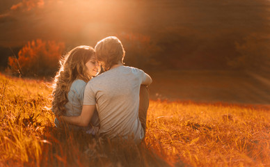 Stylish young couple sitting on a hill and admiring the sunset. A film photo with a light and a sunlight, a foreshortening from the back. Enamored youth in the second before the kiss - obrazy, fototapety, plakaty