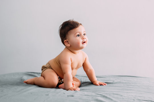 Front view of beautiful smiling baby sitting on the bed in the room, wearing shorts, family lifestyle