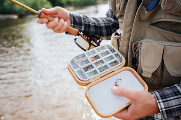 Close up of wooden box with diffeent kind of artificial silicone fishing flies and baits in it. Man holds it in one hand and has rod in the other one. He wears vest and standing in water.