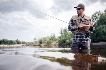 Cool fisherman stands in water and looks straight forward. He is serious. Guy holds fly rod and...