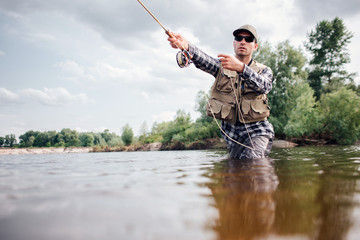 Fisherman in action. Guy is throwing spoon of fly rod in water and holding part of it in hand. He looks straight forward. Man wears special protection clothes.