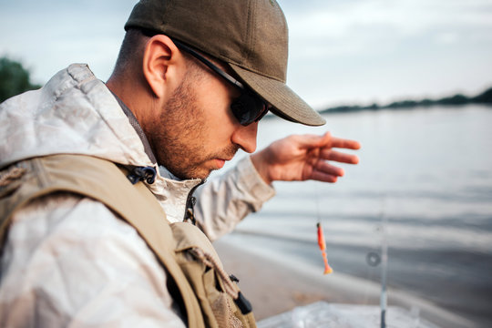 A picture of man sitting at the edge of water and looking down. He holds spoon with bait. Guy is preparing to fishing.