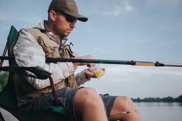 Obraz premium A picture of man sitting at the edge of water in folding chair and holding fly rod. He has small plastic bag with artificial baits in it. He reaches one of them.