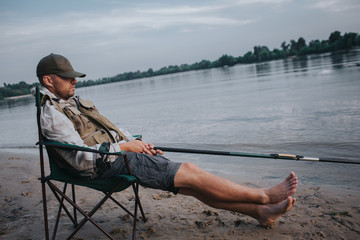 Fisherman is sitting in folding chair and sleeping. His cap is lying on eyes. Man holds fly rod in hands. He is fishing. It is evening outside.
